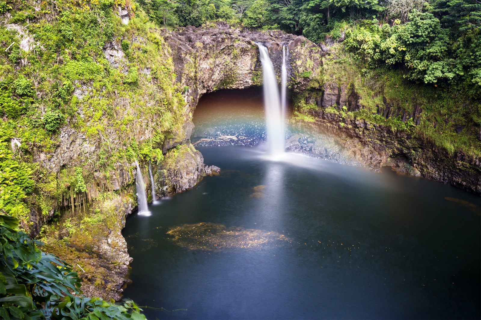 Rainbow Falls in Hilo pouring into a dark pool, with a faint rainbow in the mist and green rain forest on the cliffs