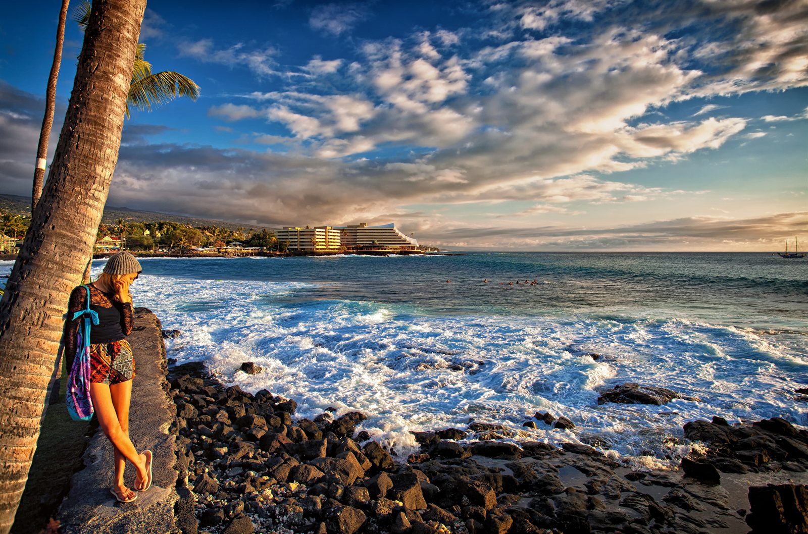 A visitor leaning against a palm trunk above the rocky lava shoreline of Kailua-Kona, with waves breaking and a hotel on the point beyond