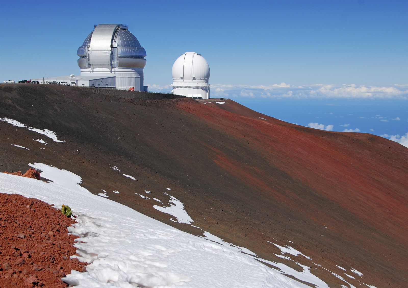 Two white observatory domes on the reddish cinder slopes of Maunakea, with patches of snow and clouds far below